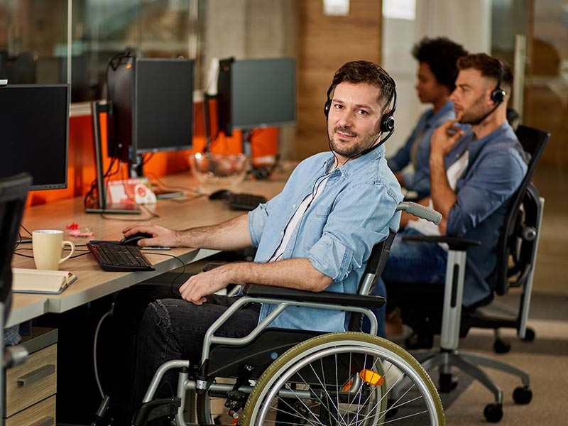 A young man in a blue shirt and black paints sits in a wheelchair at a computer workstation. Other coworkers sit at workstations nearby. ©istockphoto.com