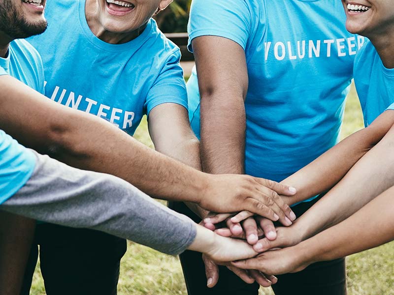 A group of five people wear light blue shirts with the word volunteer printed on the front in white letters; they all have their hands joined in the middle of the group.
