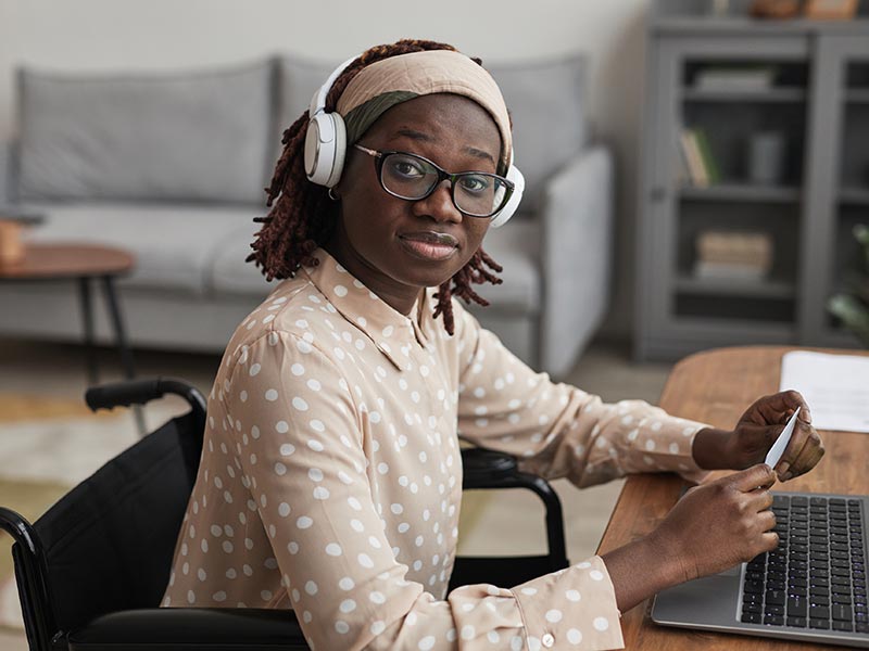 A woman wearing glasses and headphones sits in a wheelchair. She is working on laptop at a desk. ©istockphoto.com