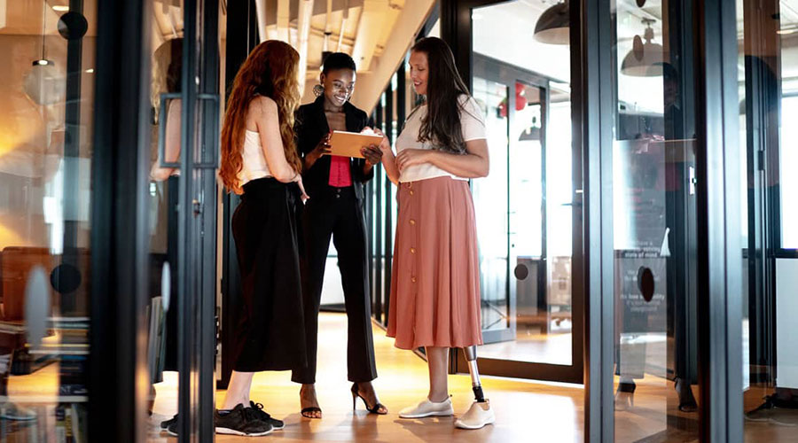 Three women smiling and looking at a computer tablet while standing in the doorway of an office.